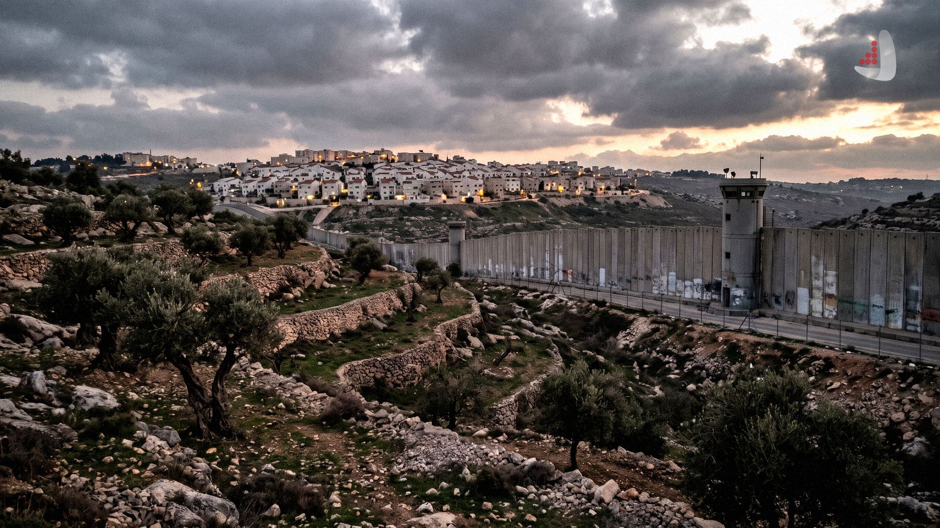 Concrete separation wall and military watchtower bordering terraced olive groves and a dense settlement