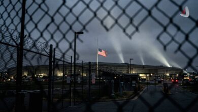 The Pentagon building at night with searchlights and the US flag at half-mast viewed through a fence