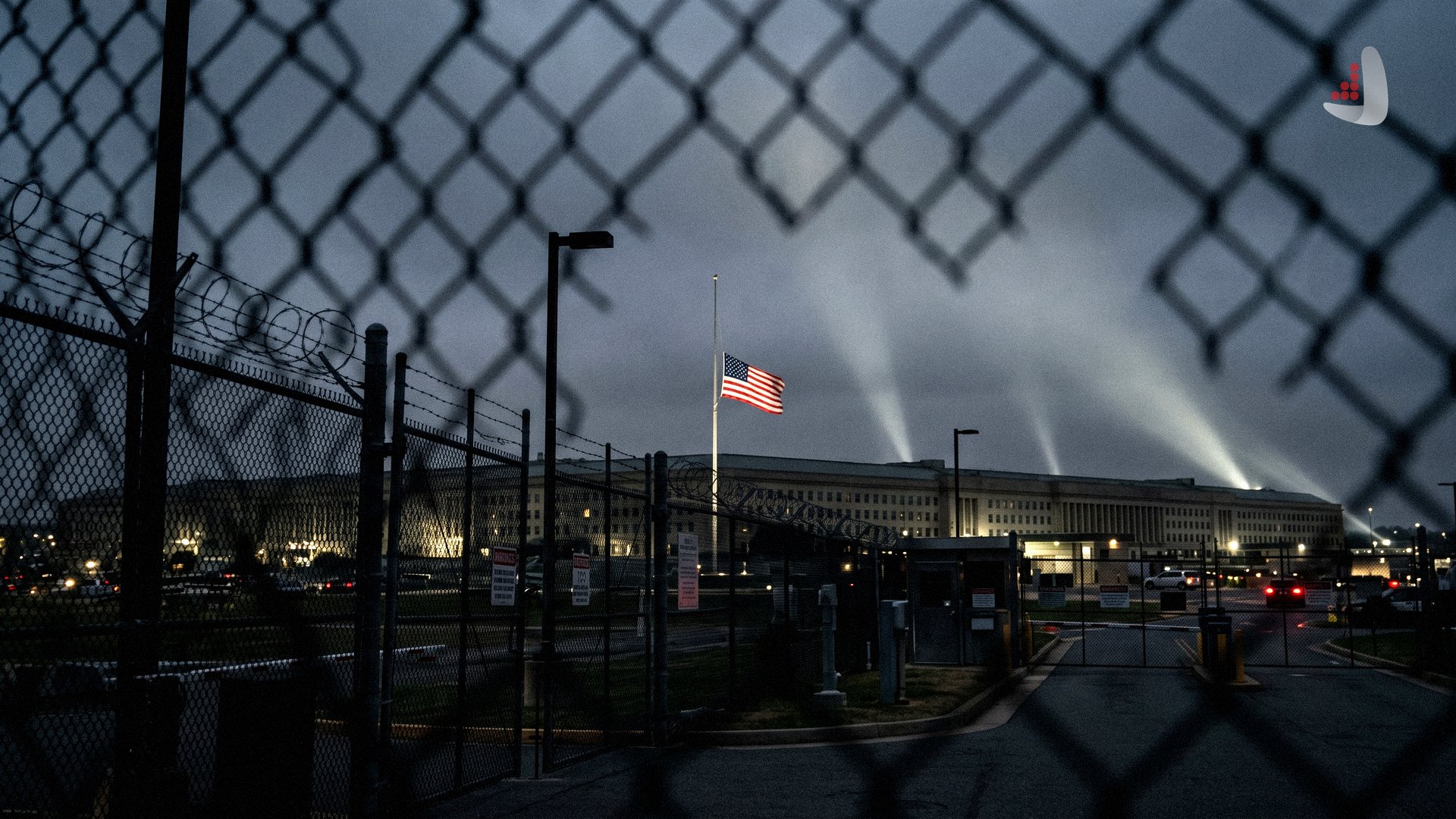 The Pentagon building at night with searchlights and the US flag at half-mast viewed through a fence