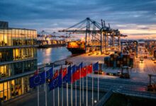Flags of the European Union and China flying at a busy industrial shipping port at sunset