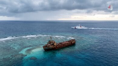 A rusted grounded ship on a reef with a white coast guard vessel passing nearby