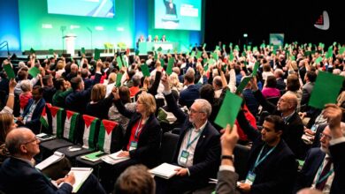 Audience members raising green voting cards at a political conference with Palestinian flags on chairs