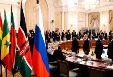 Vladimir Putin shaking hands with an African leader at a large round conference table surrounded by national flags