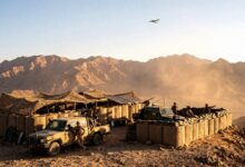 Military soldiers and pickup trucks stationed at a sandbagged outpost in a mountainous desert region