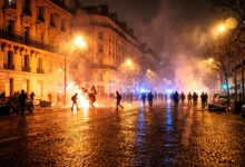 Protesters marching through Paris streets amidst street fires, smoke, and riot police presence