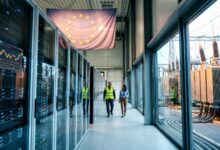 Two people walking in a data center corridor alongside server racks and electrical transformers